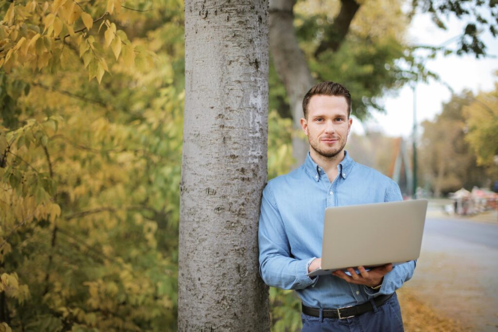 Business owner leaning on a tree while holding a laptop